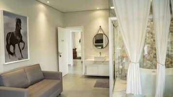 Une salle de bain moderne avec une douche, un lavabo et un miroir. On y trouve un rideau blanc, un canapé beige et un tableau encadré au mur.