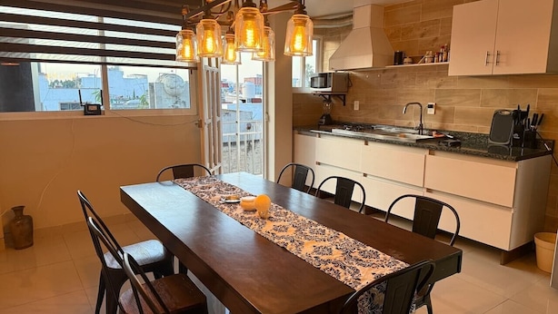 A dining area with a long table, chairs, a chandelier, a kitchen with cabinets, and a view through a window.