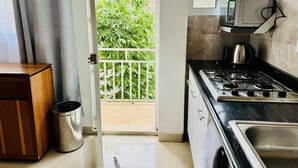 A kitchen with a gas stove, a microwave, and a sink. There is a trash can and a wooden cabinet. A door leads to a balcony with a view of greenery.