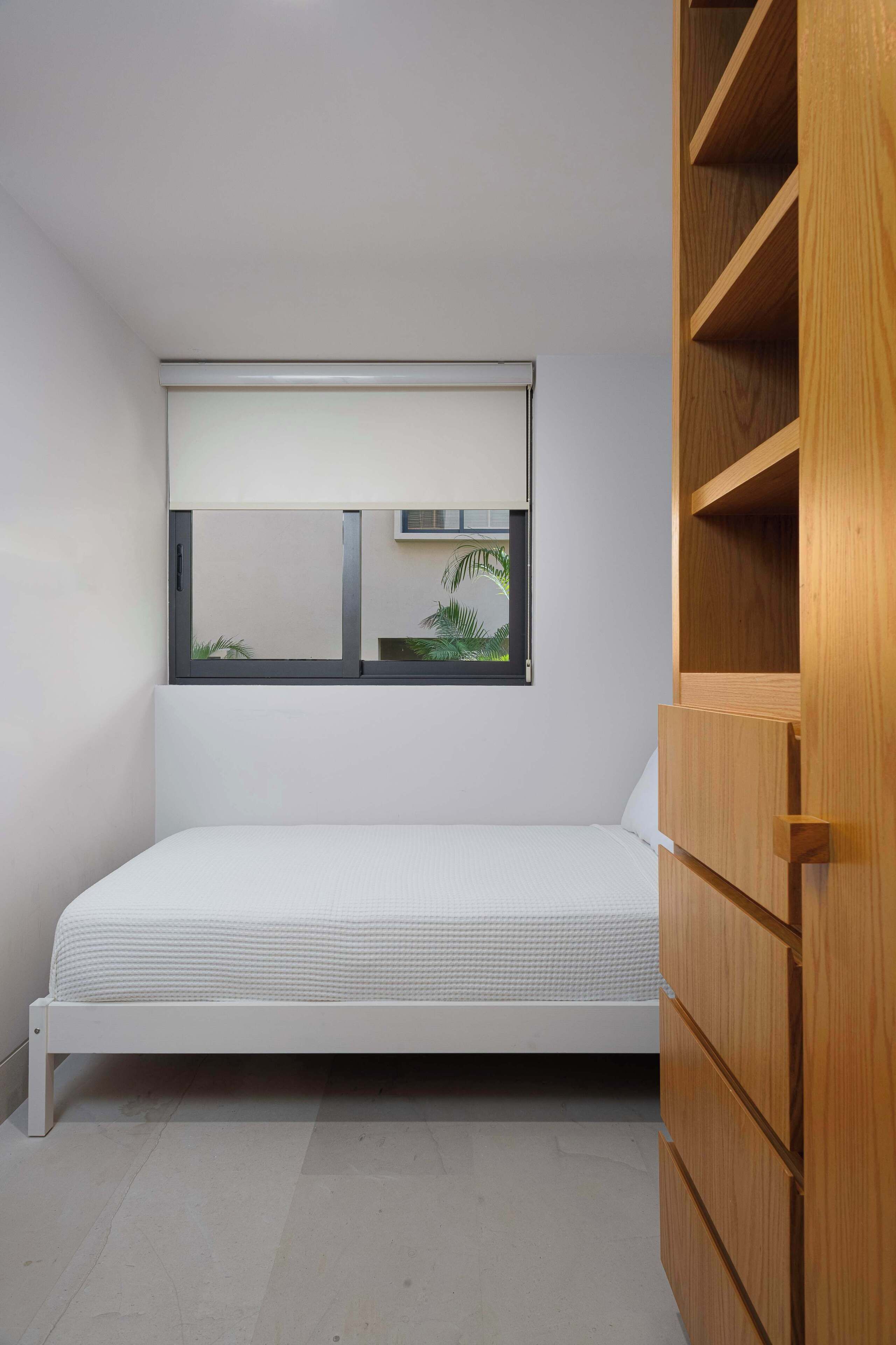 Habitació petita amb un llit individual, una finestra, un armari de fusta i un matalàs blanc.