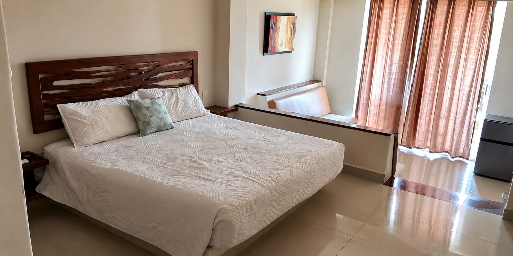 Una cama bien tendida con cabecera de madera, una mesita de noche, una silla y una ventana con cortinas.