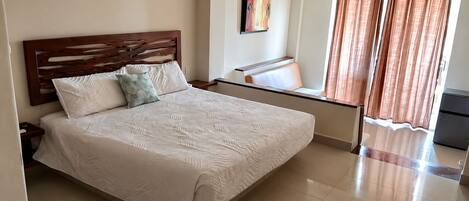Una cama bien tendida con cabecera de madera, una mesita de noche, una silla y una ventana con cortinas.