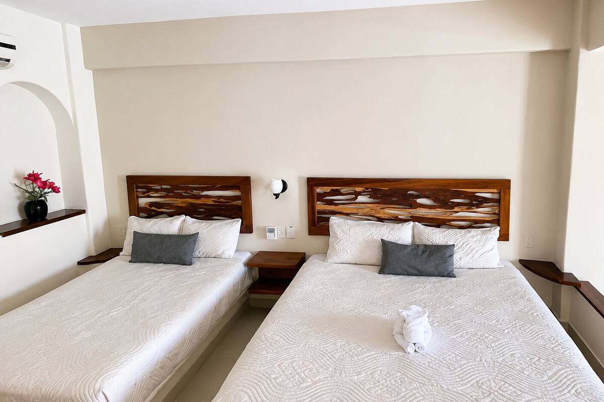 Habitación de hotel con dos camas, cabeceros de madera, ropa de cama blanca, una mesita de noche y una pequeña mesa.