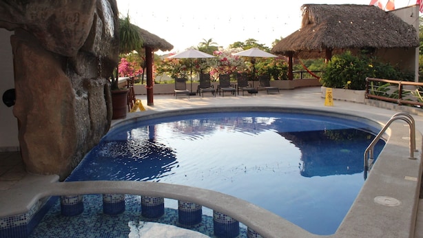 A swimming pool with lounge chairs and umbrellas, surrounded by tropical plants and a thatched-roof structure.