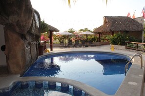 Una piscina con sillas de descanso y sombrillas, rodeada de plantas tropicales y una estructura con techo de paja.