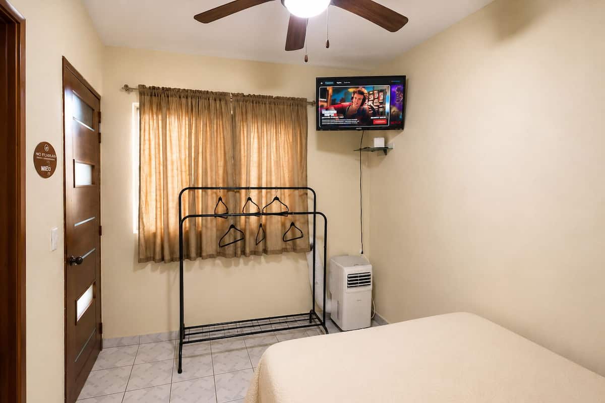 Habitación de hotel con cama, televisión montada en la pared, ventana con cortinas, radiador y puerta con cartel.