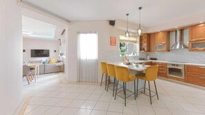 Een moderne keuken met houten kastjes, een eethoek met gele stoelen, en een televisie in de aangrenzende woonkamer.