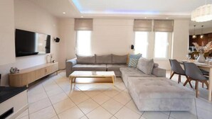 Een moderne woonkamer met een hoekbank, een salontafel, een aan de muur bevestigde televisie, en een eethoek met stoelen en een tafel.