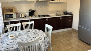 A kitchen with a microwave, stove, sink, and a refrigerator. There is a dining table with chairs and a patterned tablecloth.