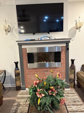 Ein modernes Wohnzimmer mit einem flachbildfernseher über einem Kamin, einer dekorativen Vase und einer Blumenkomposition auf einem Teppich.
