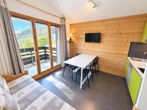 A room with a large sliding glass door leading to a balcony, a wooden wall, a flat-screen TV mounted above a white table with chairs, and a kitchen area with green cabinets. - 10m from ski slopes, 4th floor, view valley, balcony, tv, ski locker, 24m², Les Menuires (Les Menuires)