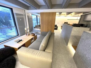 Ein modernes Wohnzimmer mit einer Couch, einem Holztisch mit Schachfiguren und Blick auf eine verschneite Landschaft durch große Fenster.