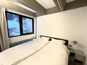 Ein Schlafzimmer mit einem Bett, einem Nachttisch mit Lampe, einem Fenster mit Vorhängen und Blick auf eine verschneite Landschaft.