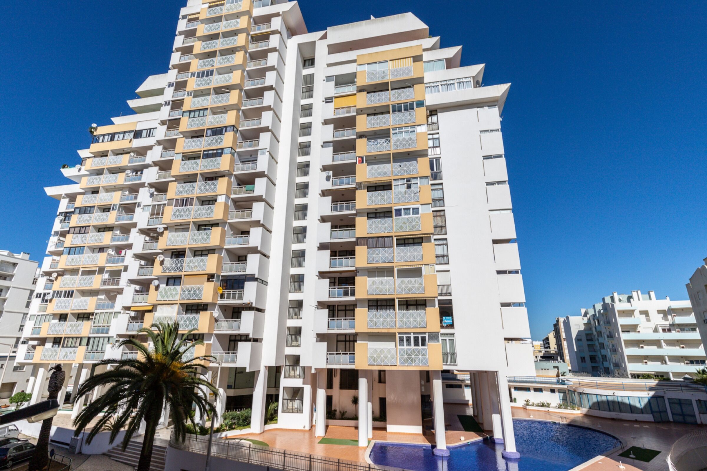A multi-story hotel with balconies, a swimming pool, and a palm tree.