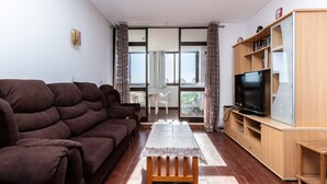 A living room with a sofa, a TV on a wooden stand, a table with a tablecloth, and a view through a glass door. - Sagan Brown Apartment, Armação de Pêra, Algarve (Armação de Pêra)