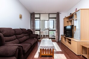 Ein Wohnzimmer mit einer Couch, einem Fernseher auf einem Holzboden, einem Tisch mit Tischdecke und Blick durch eine Glastür.