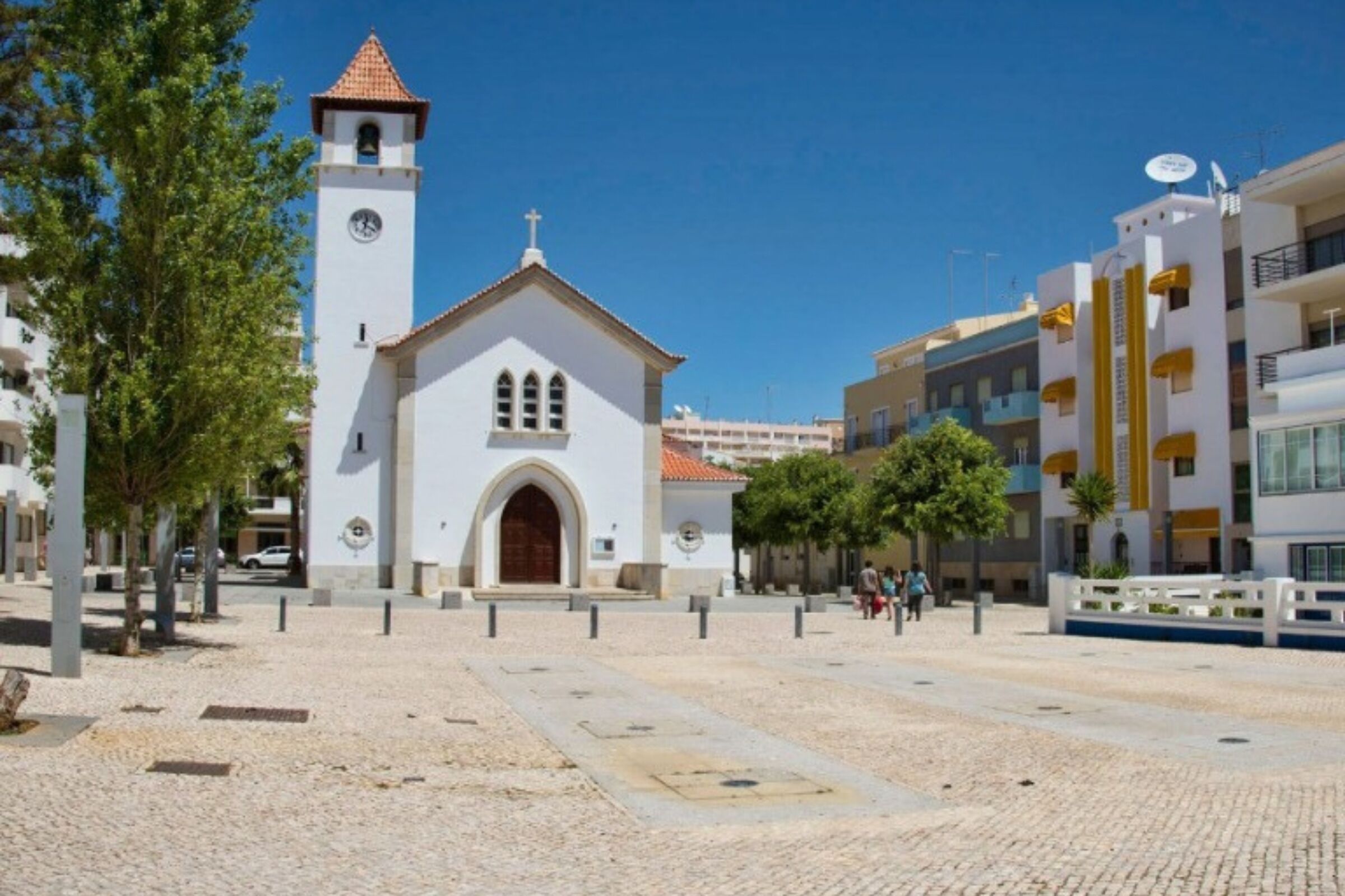 Uma igreja com uma torre do relógio, calçada de pedra e edifícios modernos ao fundo.