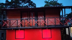 Un edificio con fachada roja, un balcón con barandilla de metal y plantas en macetas a nivel de calle.
