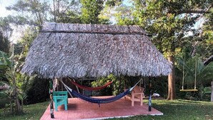 Una estructura de techo de paja con una hamaca, un banco y una pequeña mesa, rodeada de hierba y árboles.