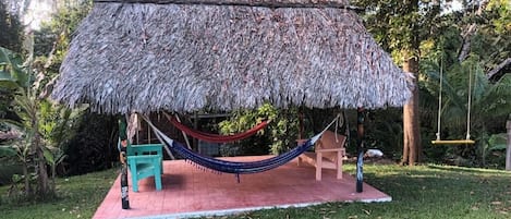 A thatched-roof structure with a hammock, a bench, and a small table, surrounded by grass and trees.