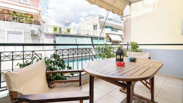 A balcony with a table and chairs, a clock, and a view of a swimming pool and buildings. - Casa Athenia Inn (Athina)