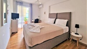 A modern hotel room with a large bed, a desk, a chair, a TV, and a small table with a lamp. - Casa Athenia Inn (Athina)