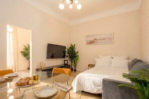 Ein modernes Hotelzimmer mit einem Bett, einem Fernseher, einem Esstisch mit Tellern und Brot sowie zwei Topfpflanzen.