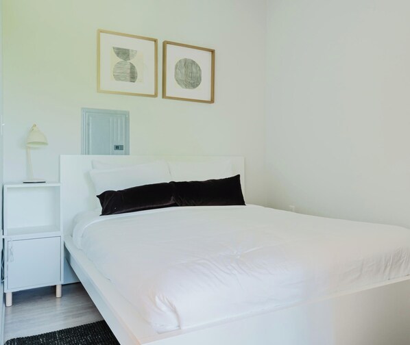A neatly made bed with white bedding, a white nightstand with a lamp, and two framed artworks on the wall. - Joinery North (Charlotte)