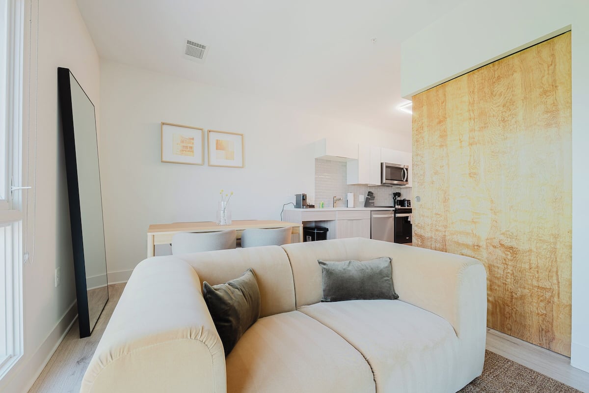 Una sala moderna con un sofá color beige, un televisor de pantalla plana y una cocina al fondo.