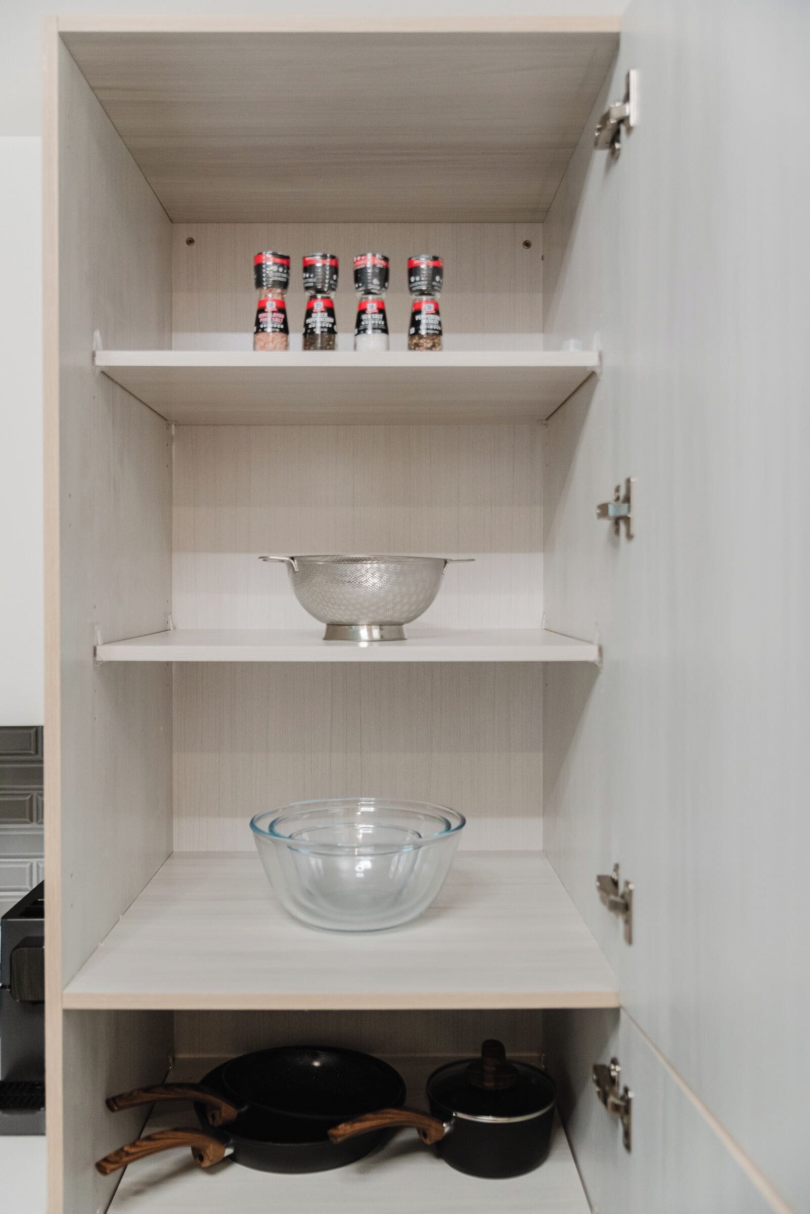 A kitchen cabinet with shelves, a colander, a glass bowl, and a frying pan.
