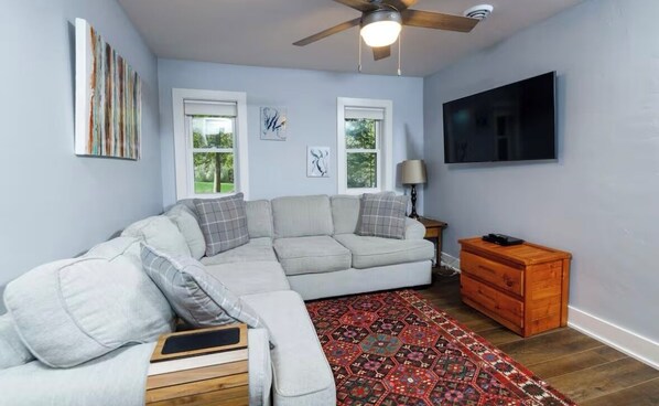Ein Wohnzimmer mit Ecksofa, einer Holzkiste, einem gemusterten Teppich und einem Deckenventilator.