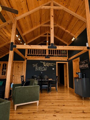 Ein rustikaler Innenraum mit freiliegenden Holzbalken, einem Essbereich und einem Schild mit der Aufschrift 'THE BURROW COFFEE BAR'.