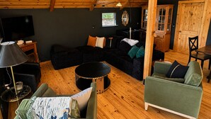 Un salon chaleureux avec un plafond en bois, un canapé noir, un fauteuil vert, une table basse ronde et un téléviseur.