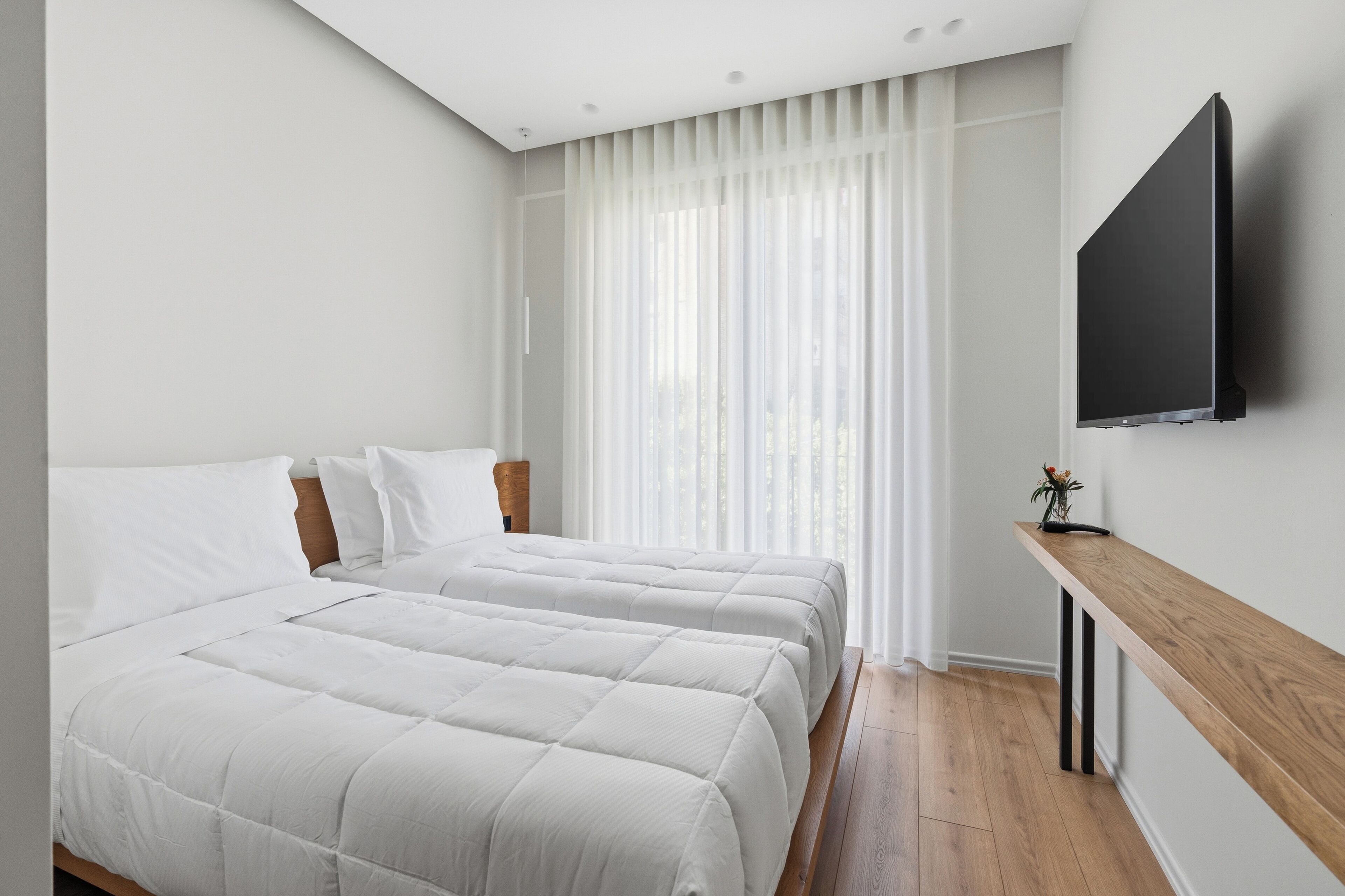 A bedroom with two beds, a TV mounted on the wall, and a window with sheer curtains.