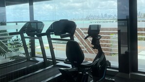 A gym with exercise equipment and a view of a city skyline through large windows. - Restinga 11 · Beachfront apartment in Caribessa with private balcony (João Pessoa)