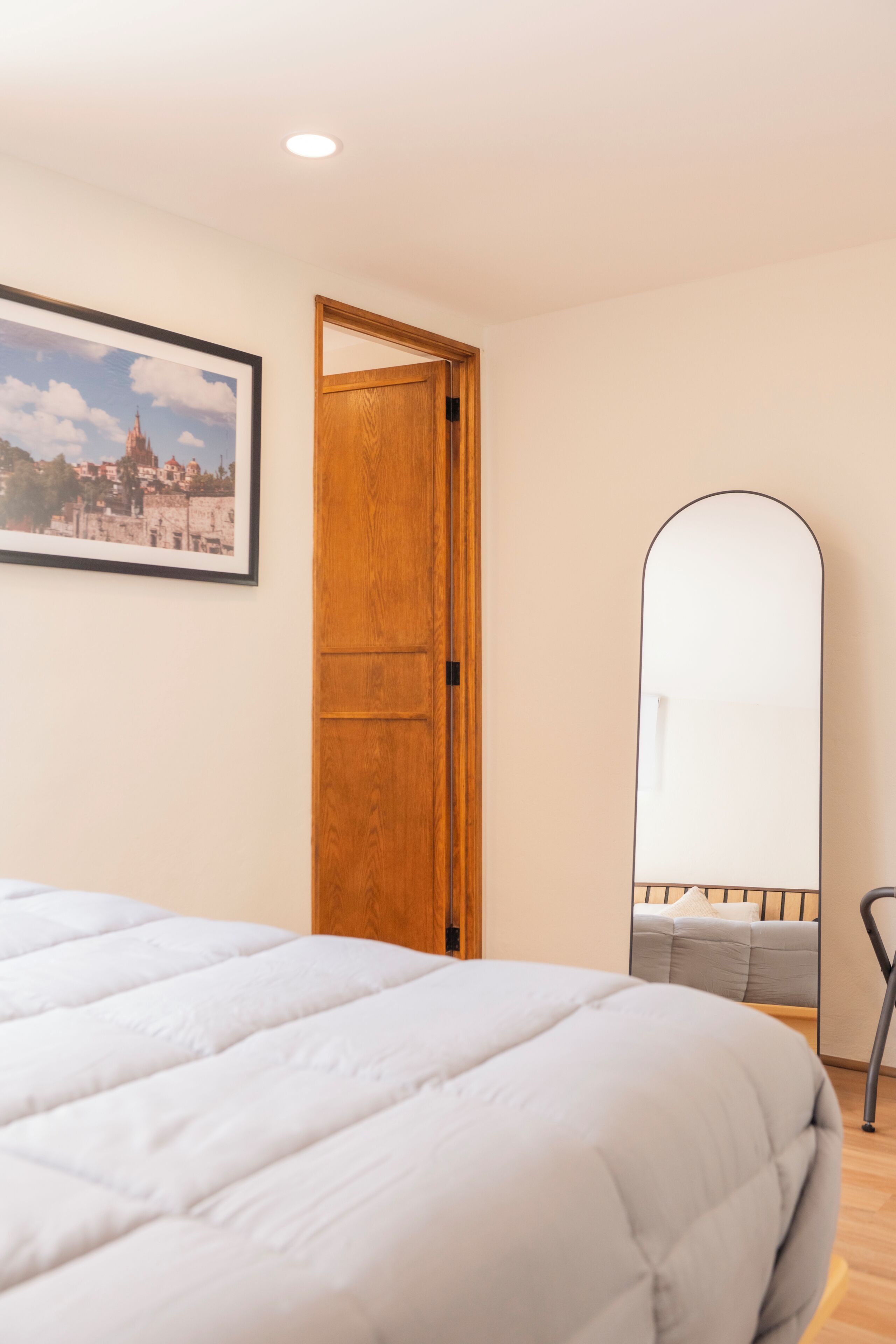 Un dormitorio con cama, una puerta de madera, un cuadro enmarco y un espejo.
