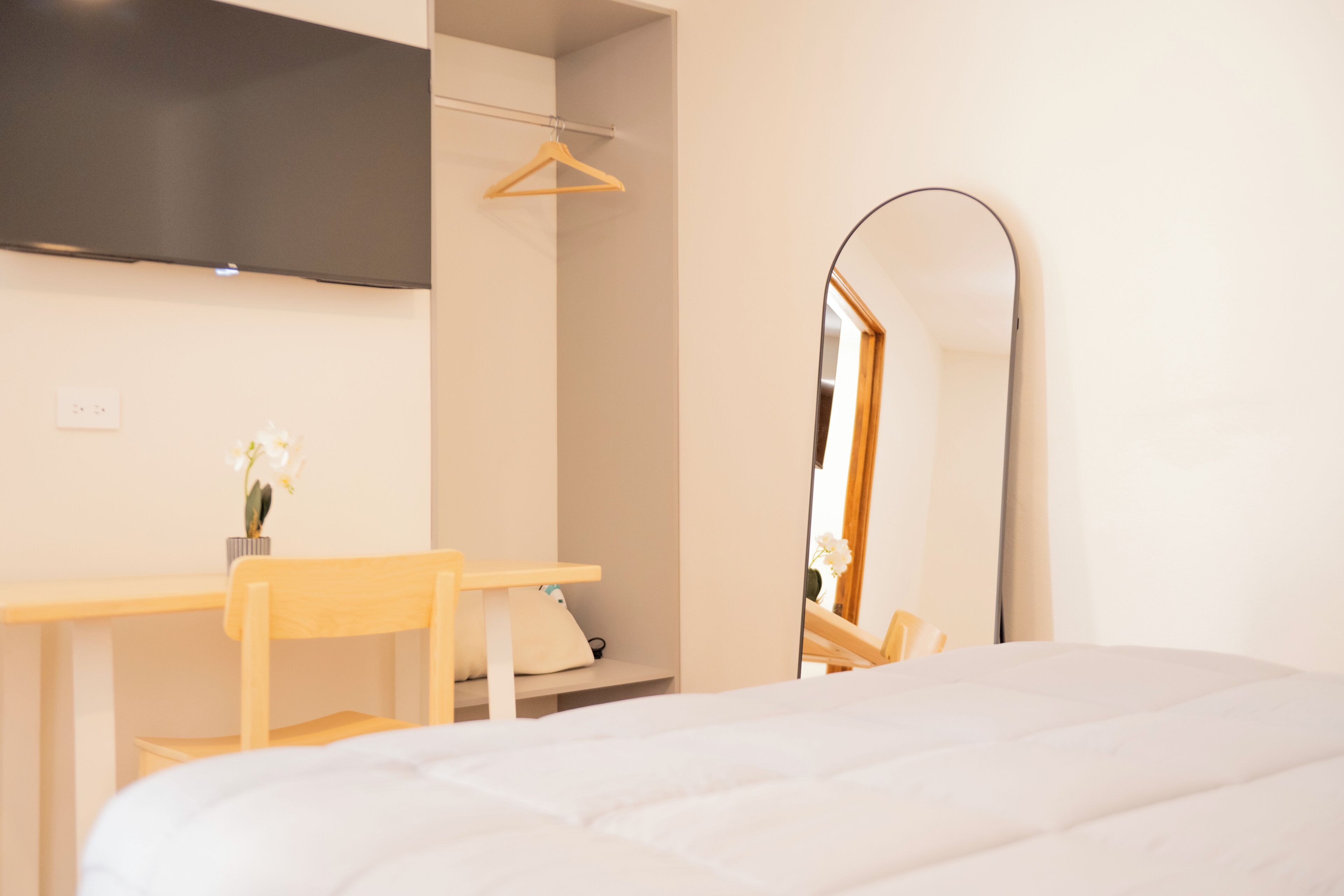 A bedroom with a bed, a desk, a chair, a TV mounted on the wall, and a mirror.