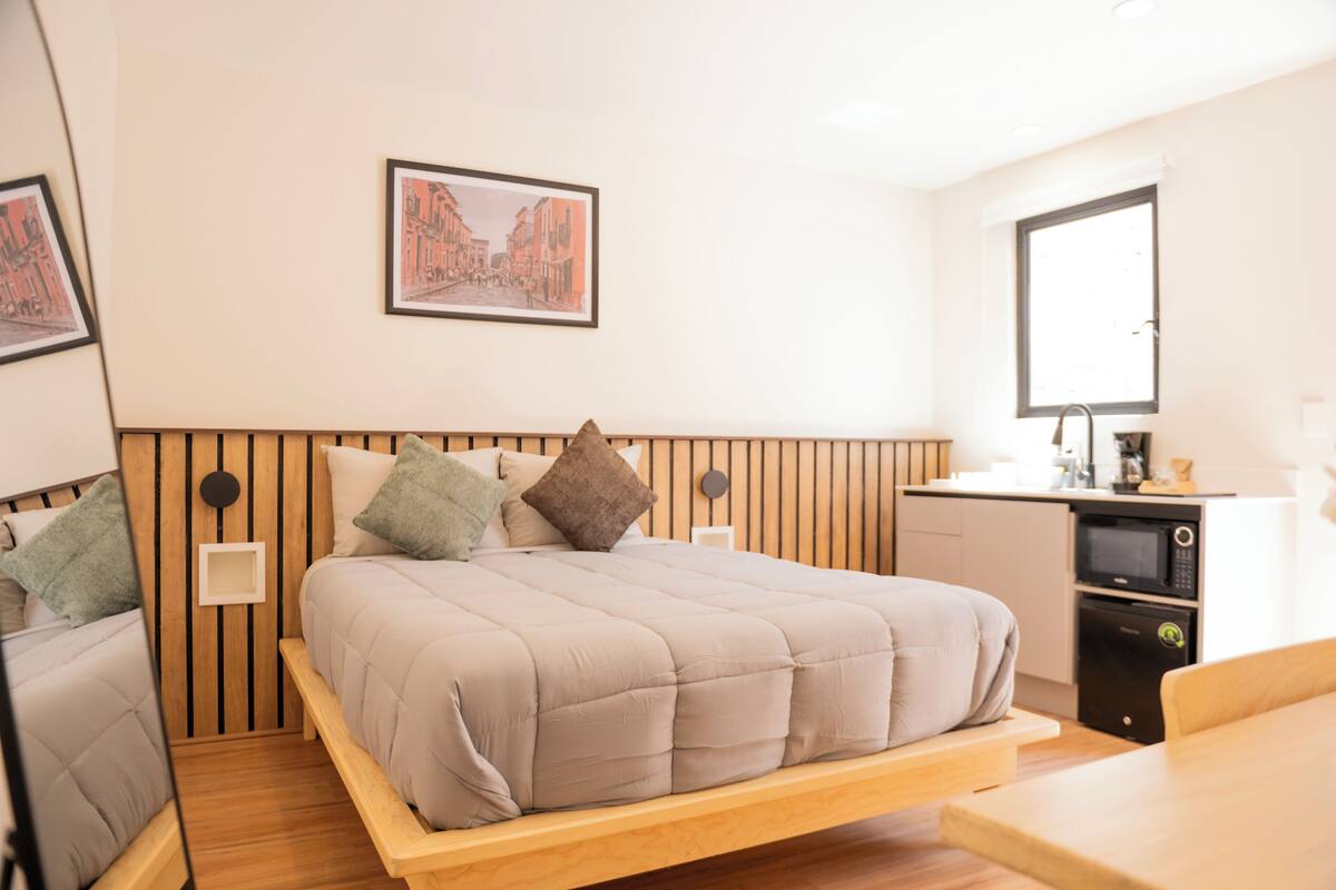 Un dormitorio con cama, mesitas de noche, televisor en la pared, ventana y kitchenette.