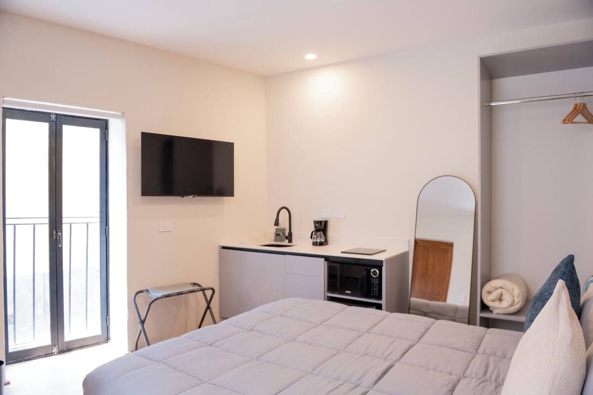 Un dormitorio con cama, televisor montado en la pared, escritorio con cafetera, microondas y espejo.