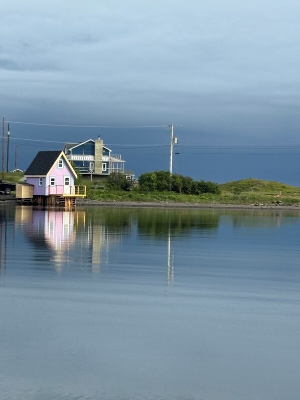 Une maison rose avec un balcon, une maison plus grande en arrière-plan, et une étendue d’eau calme qui reflète les bâtiments.