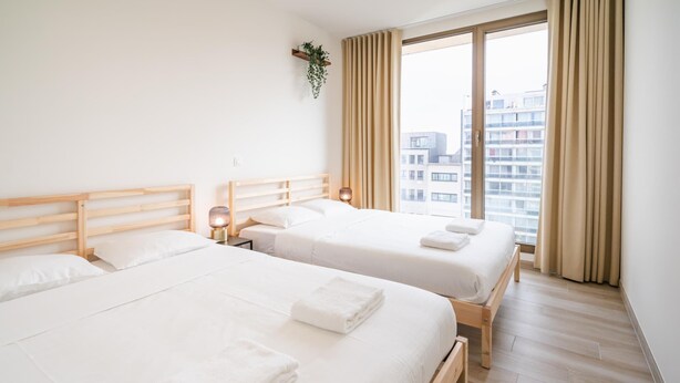 Hotellihuone, jossa on kaksi sänkyä, puinen pääty, yöpöytä lampun kanssa ja suuri ikkuna, jossa on verhot.