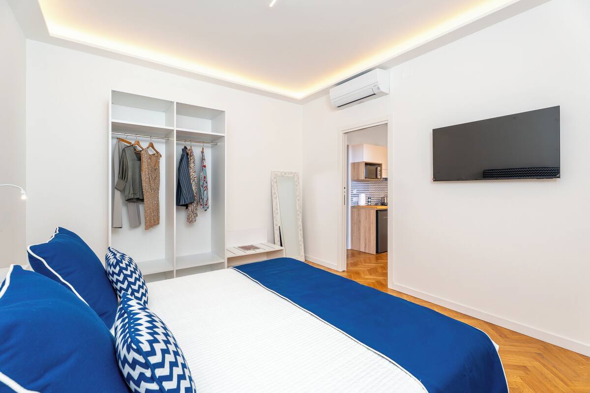 A modern bedroom with a bed, a wardrobe, a television, and a kitchenette.