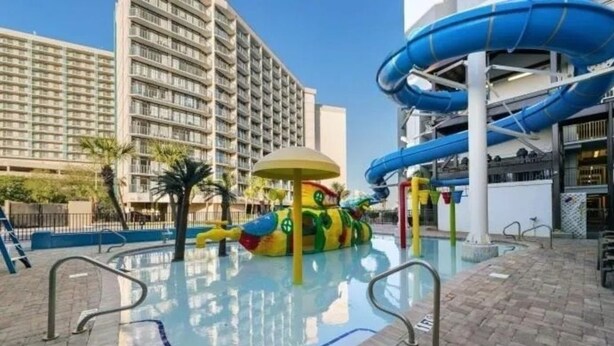 Zona de piscina con tobogán acuático, estructura de juegos para niños y un gran edificio de hotel al fondo.