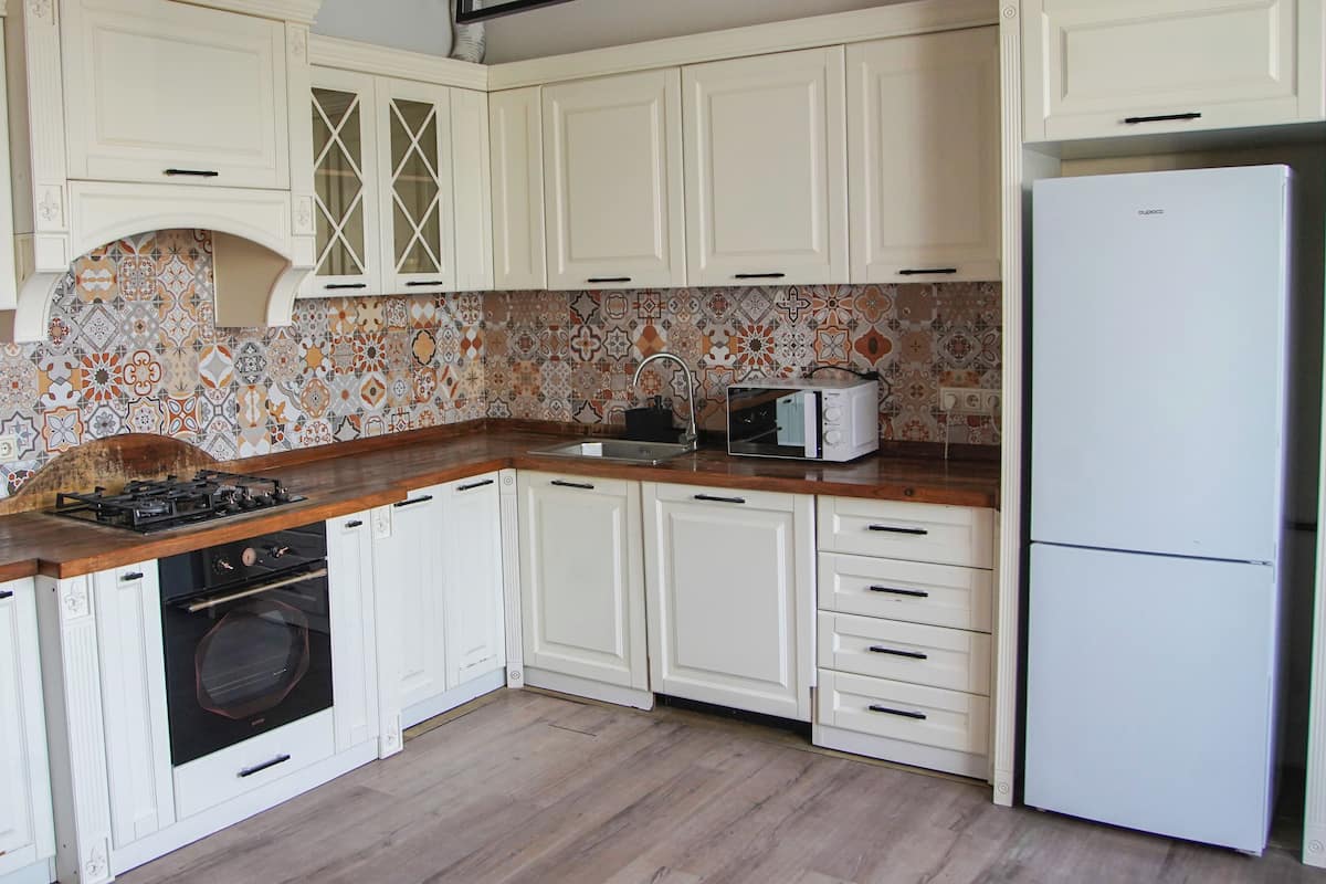 Een keuken met witte kastjes, een achterwand met patroon, een gasfornuis, een oven, een magnetron en een koelkast.