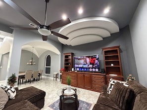Ein modernes Wohnzimmer mit Deckenventilator, Flachbildfernseher, Essbereich und einer gemütlichen Sitzecke.