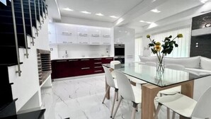 Una cocina moderna con armarios blancos, un comedor con mesa de vidrio y una escalera con barandillas negras.
