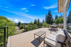 Terraza de madera con mobiliario exterior, vista a árboles y una carretera, bajo un cielo azul despejado.
