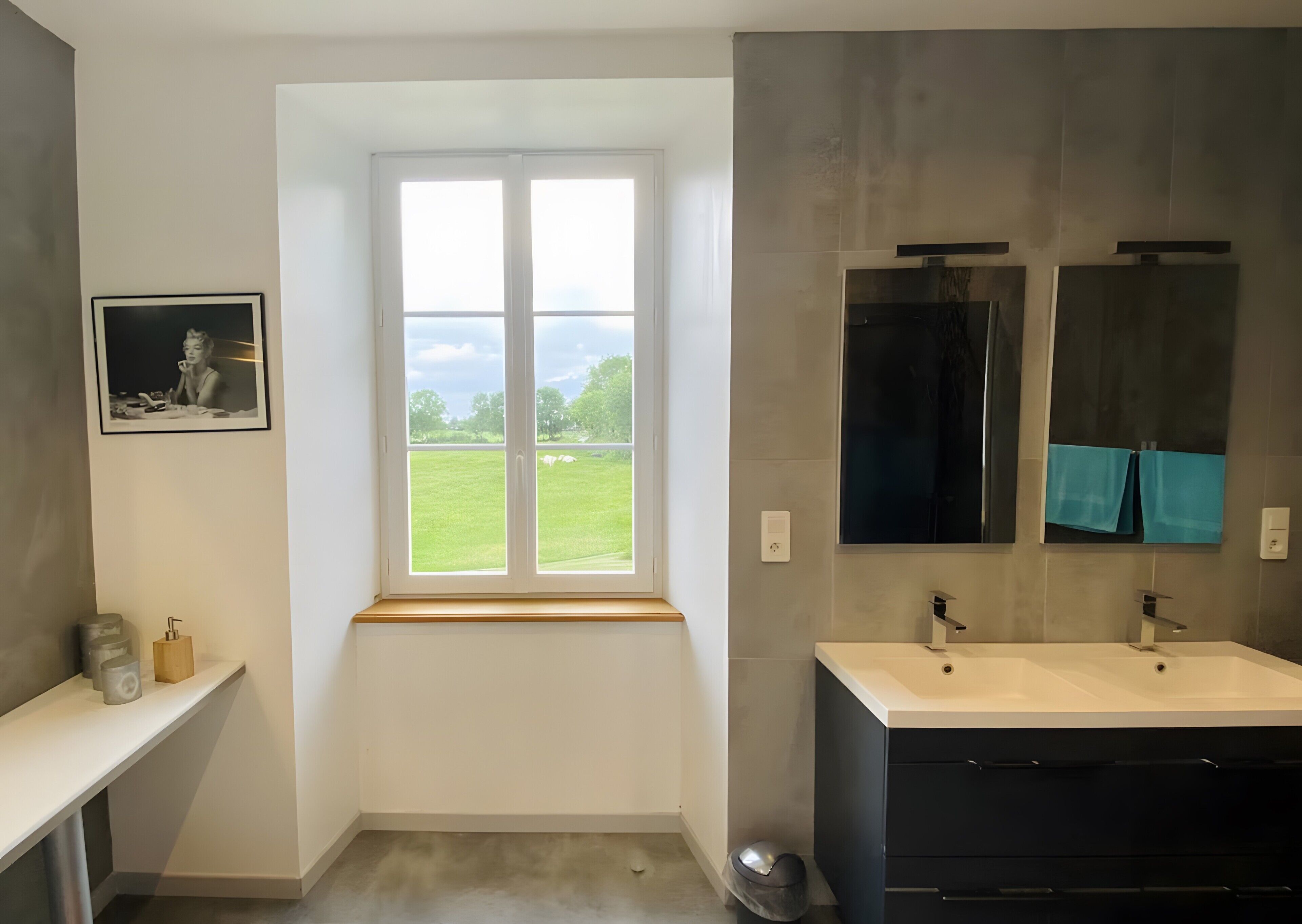 Un baño con ventana, lavamanos, espejo y una foto enmarcada en la pared.