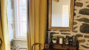 Una habitación con pared de piedra, un escritorio de madera con cajones, una silla, un espejo y una ventana con cortinas.