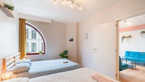 Een slaapkamer met twee bedden, een raam met gordijnen, een plant en uitzicht op een blauwe bank in de aangrenzende kamer.